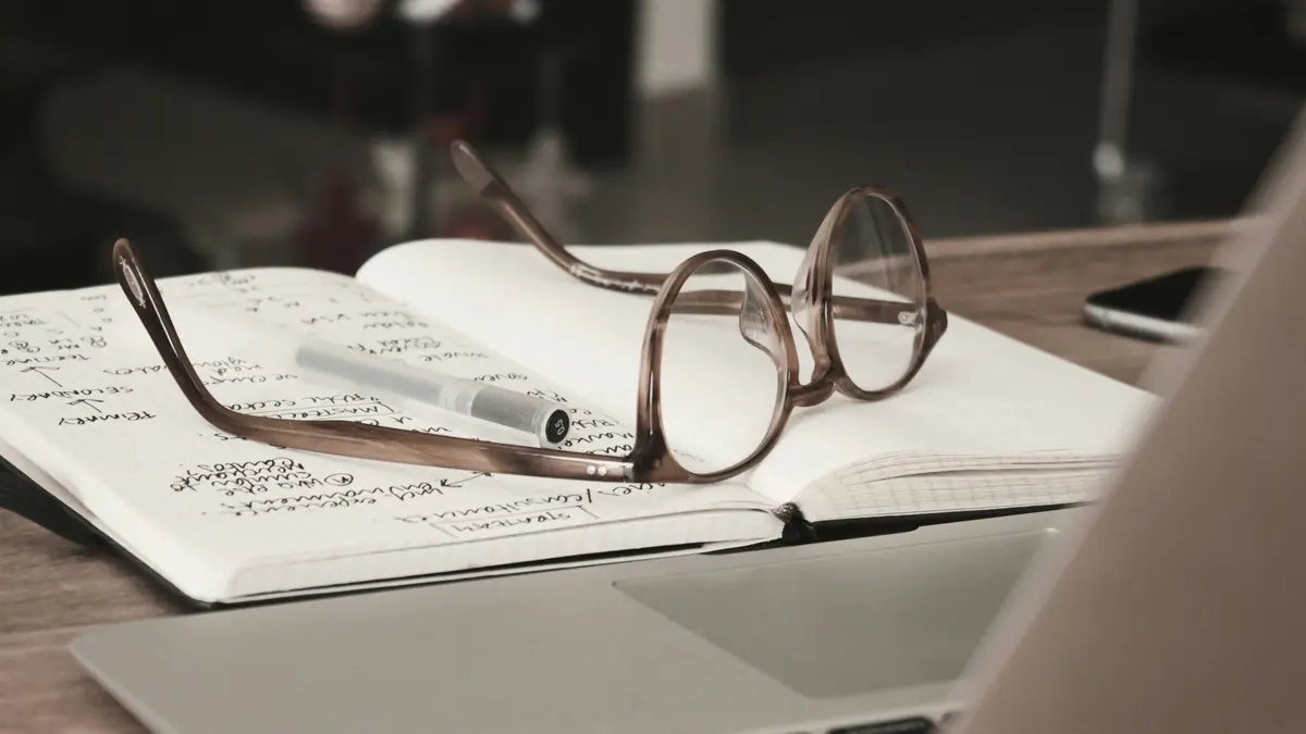 Reading glasses and pen resting on an open notebook of handwritten research notes beside a laptop